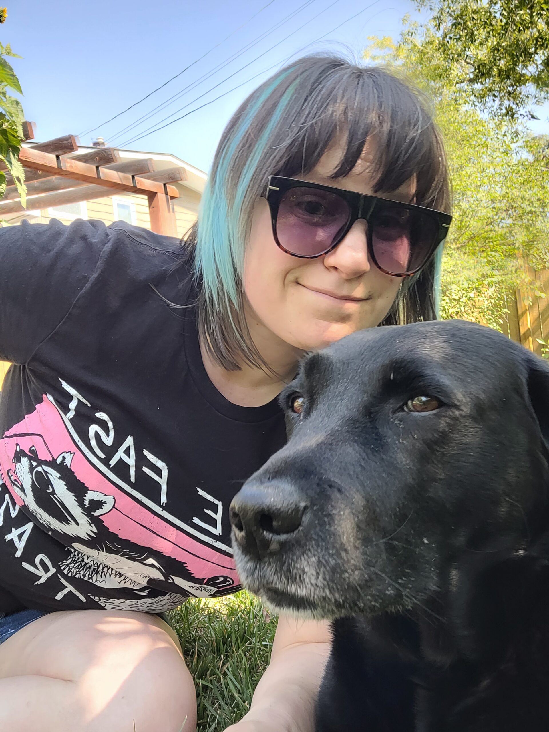 a woman posing with a dog. the woman is wearing sunglasses and is smiling. the dog is a black labrador who is looking suspiciously at the camera.